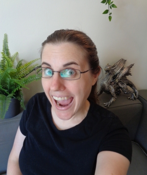 Author A. C. Spahn, a white woman with a brown ponytail and glasses, making a silly excited face, with a dragon sculpture and a fake fern in the background